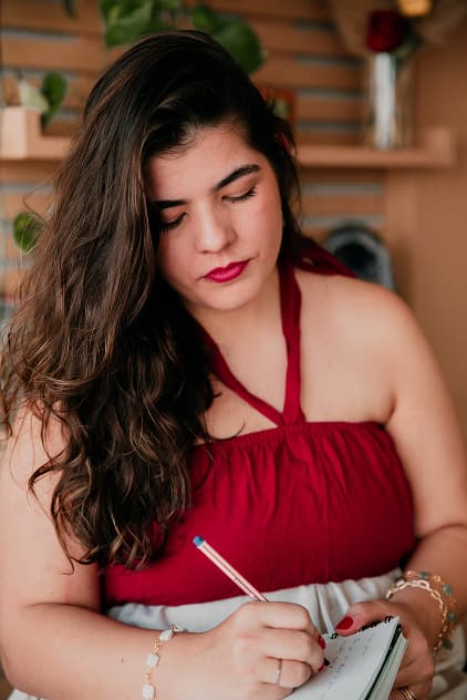 Isa Pupo em blusa vermelha com fundo com elementos naturais, concentrada anotando em um caderno.